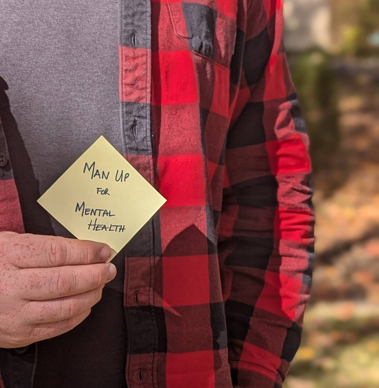 a guy wearing plaid outside holding a yellow sticky note that says "Man Up for Mental Health"